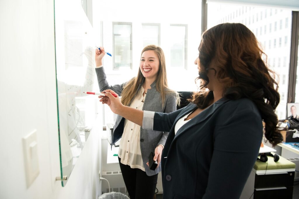 pexels-photo-1181533-1181533 Two professional women discussing ideas on a whiteboard in a modern office setting.