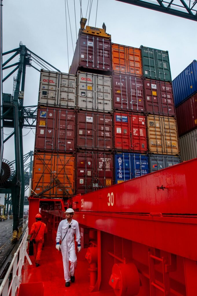 pexels-photo-28438391-28438391 Maritime workers navigating a ship deck with towering stacks of shipping containers in port.
