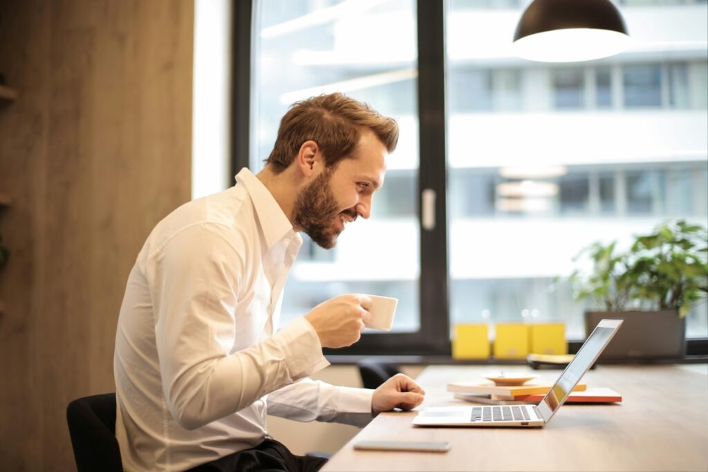 pexels-photo-925786-925786 Man in office with coffee, smiling while working at laptop, captures the essence of remote work.